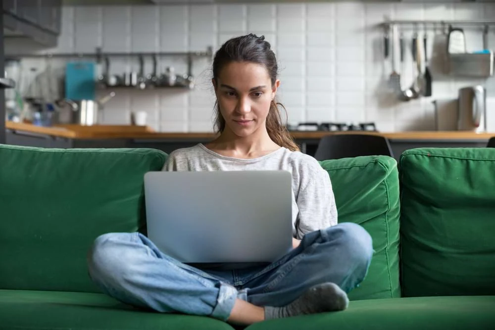 Woman using laptop on couch in modern kitchen for digital marketing agency work.
