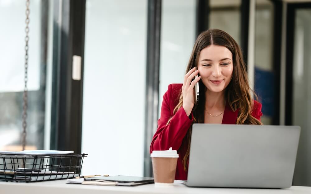 Woman in red blazer at desk using laptop and phone for digital marketing tasks.