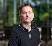 A digital marketing agency expert in a black polo stands indoors with greenery behind.