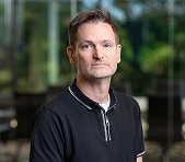 A digital marketing agency expert in a black polo stands indoors with greenery behind.