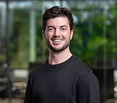 A smiling digital marketing agency professional stands indoors with greenery blurred behind.