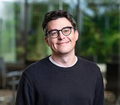 A digital marketing agency professional stands indoors with greenery outside the window.
