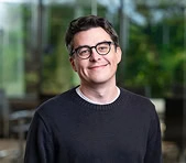 A digital marketing agency professional stands indoors with greenery outside the window.