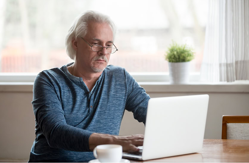 Older man uses laptop at table, researching digital marketing and SEO services.